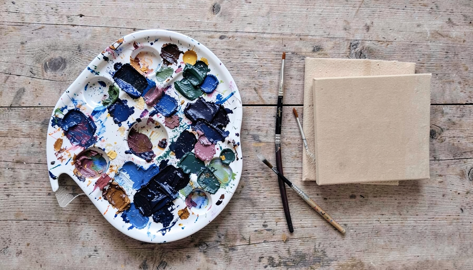 Overhead flat-lay of a painter's palette with dried oil paint, brushes, and a small stretched canvas in muted earth tones on a wooden table