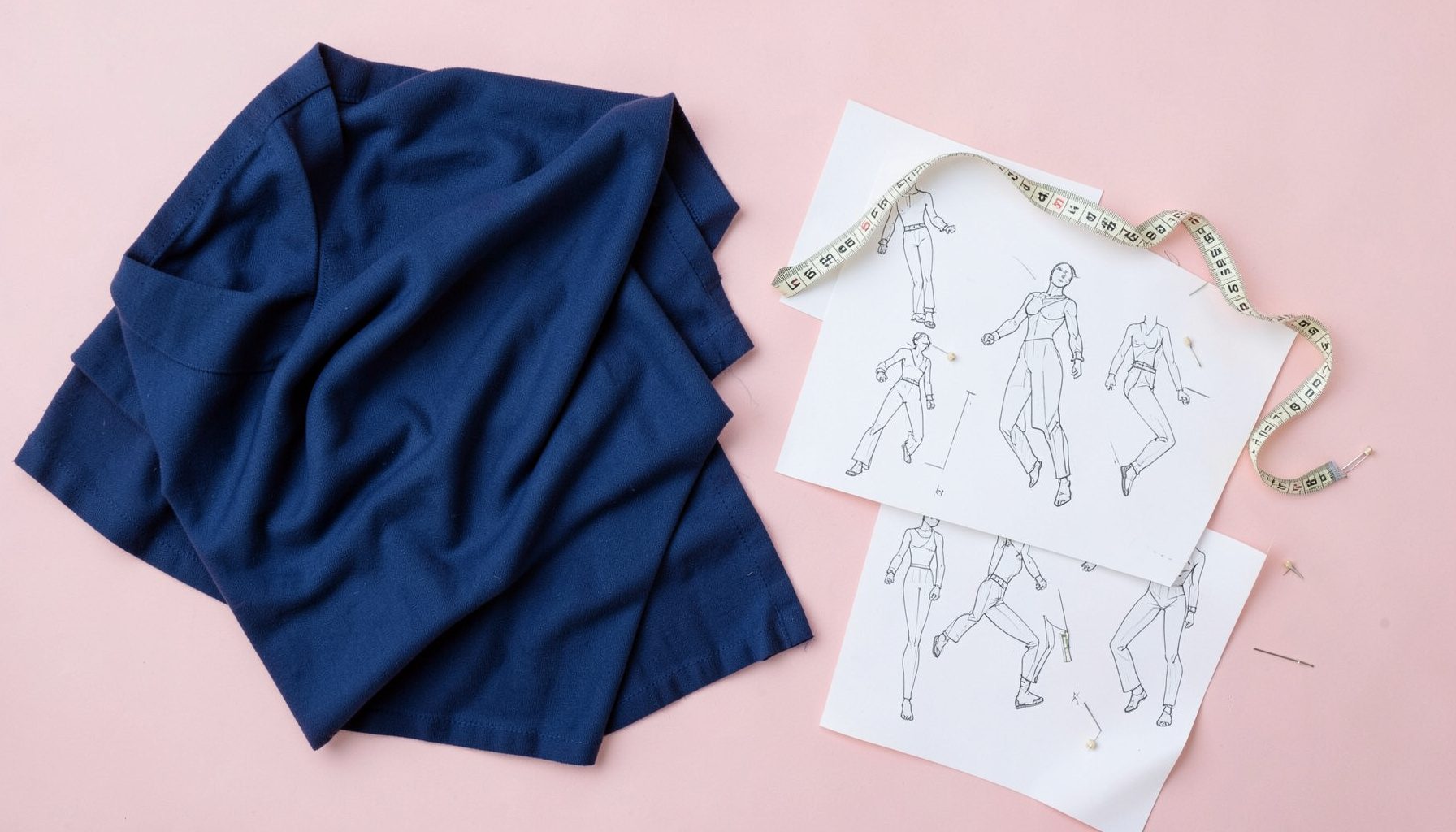 Overhead flat-lay of fashion design materials — draped fabric, fashion illustration sketches, measuring tape, and pins on a light wooden table.
