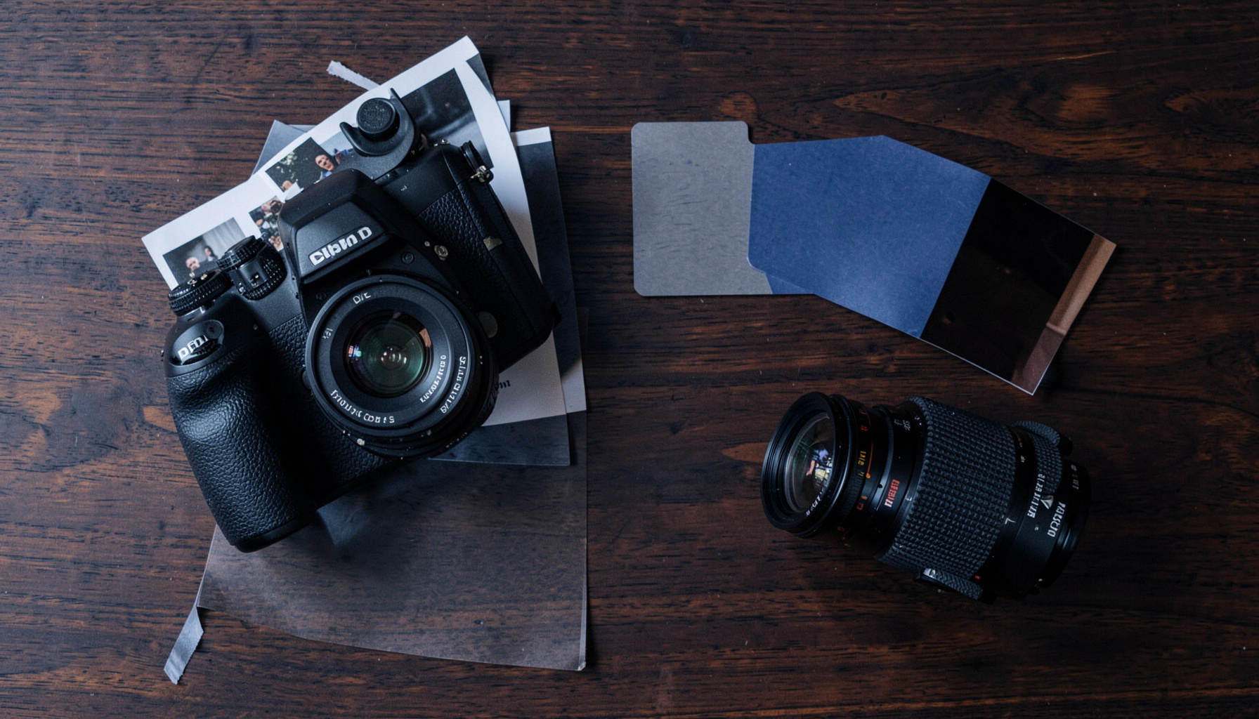 Overhead flat-lay of a DSLR camera, printed contact sheets, a grey card, and a vintage lens on a dark walnut table