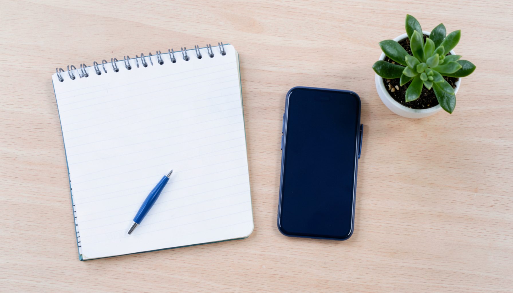 Overhead flat-lay photograph of a tidy desk with a notepad, pen, smartphone, and a small potted plant on a light wood surface.