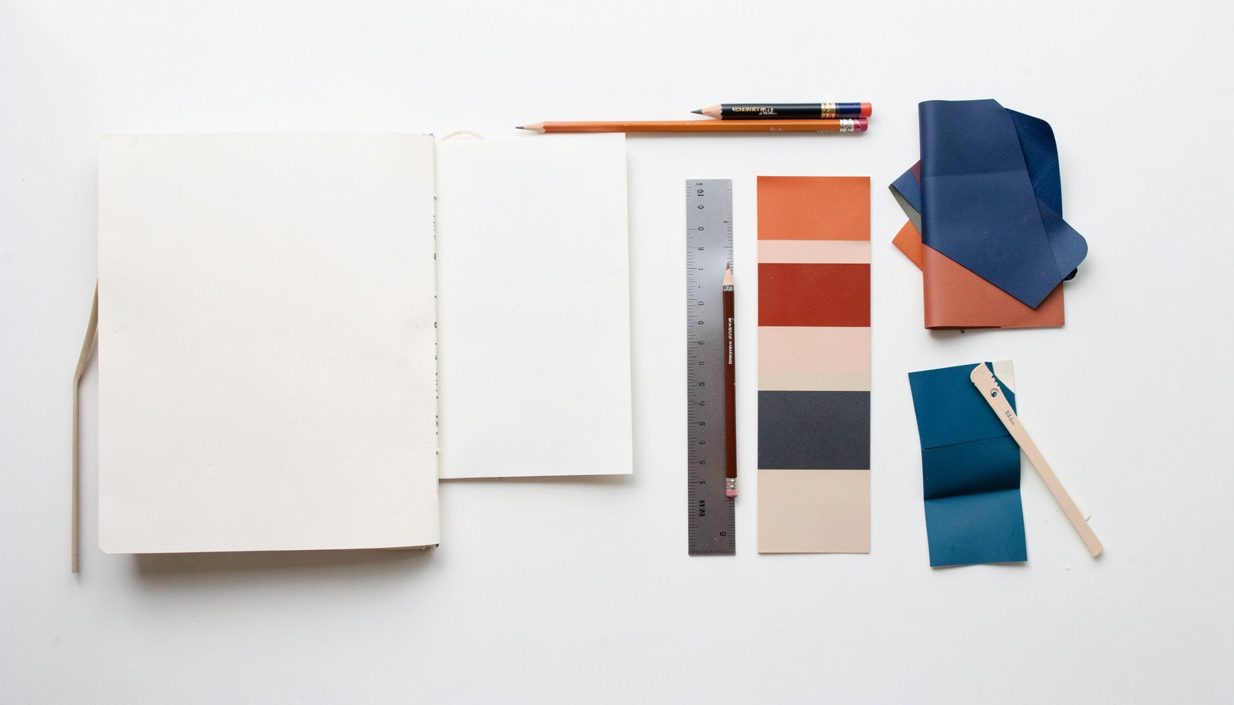 Overhead flat-lay photograph of art and design materials — sketchbook, pencils, color swatches, and a ruler — arranged neatly on a light wooden desk.